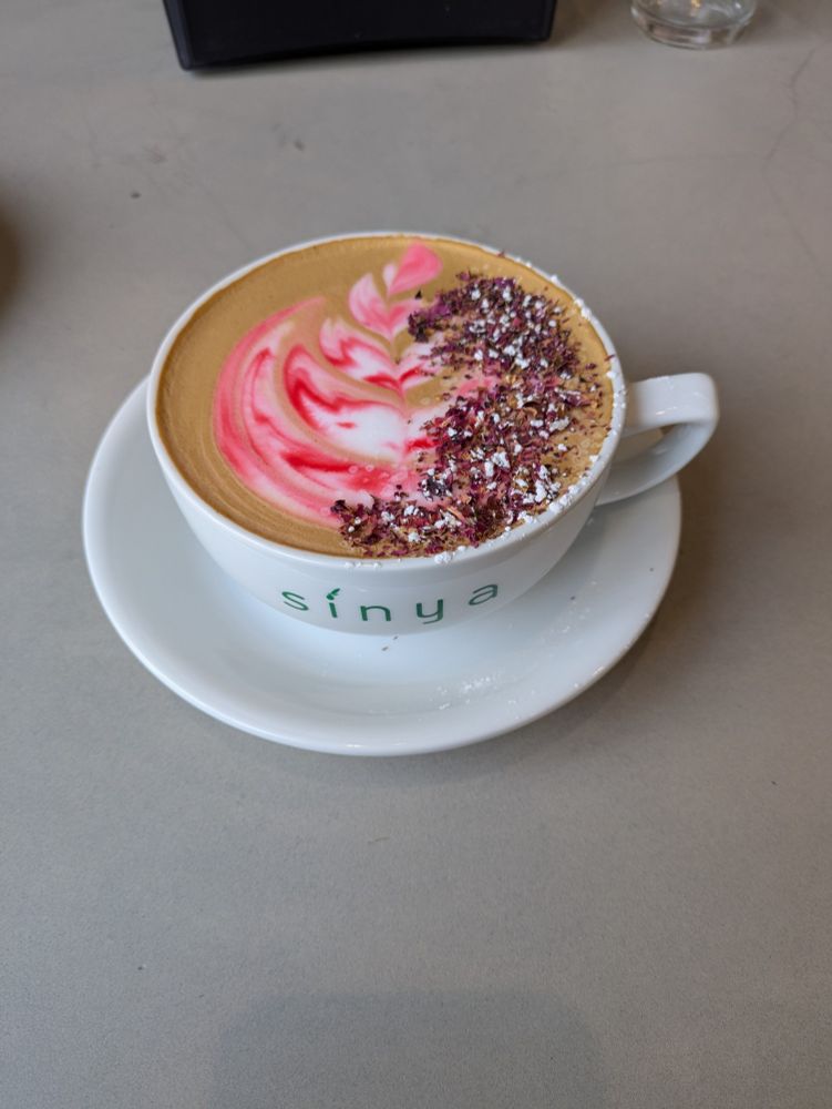 A wide coffee cup on a saucer from the restaurant Sinya. It has pink and white abstract latte art and a sprinkle of dried crushed rose petals.