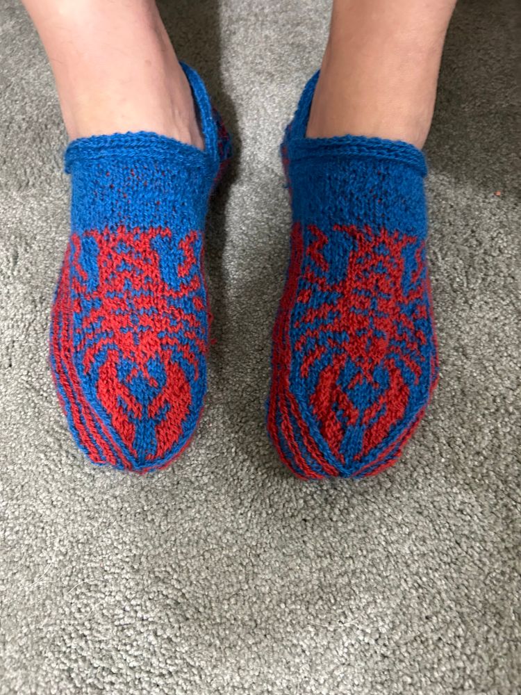 A pair of hand-knit  smucks (slippers) in red and blue yarn with a lobster pattern on the top. 