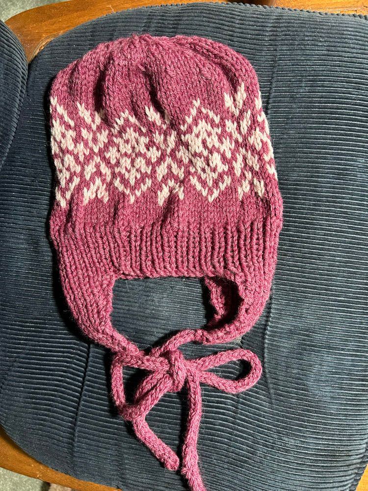 A hand-knit hat in raspberry yarn with a white color work section in a floral motif. The hat has ear flaps as part of the brim ribbing and I-cord ties. 