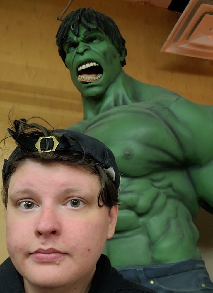 Aquillo (a white, nonbinary human) with a neutral expression wearing a black hat with messy brown hair peaking from underneath. They are wearing a black Hoodie. They stand in front of a larger than life (plastic?) figure of The Hulk whose jaw is at an unnatural angle, clearly broken. The figure is green, shirtless, wearing jeans, and has an angry expression. 