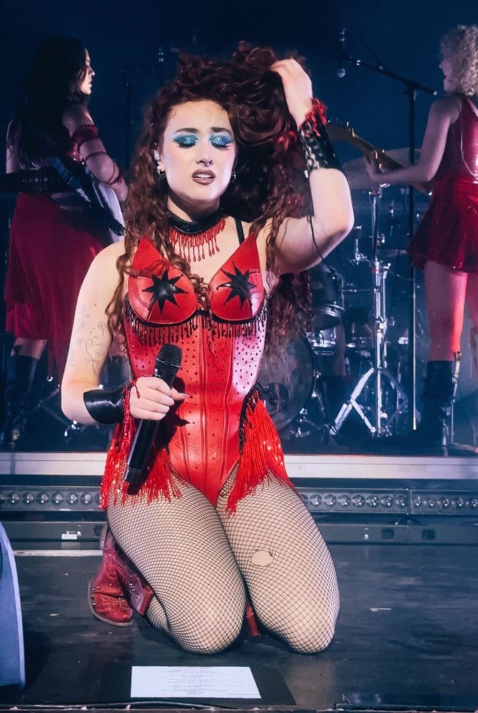 chappell roan kneels on the stage during a performance. Her face is overcome by emotions. Her eyes are closed and clad with sparkly blue eyeshadow. She is wearing a black choker with red tassels. She holds the microphone in her left hand and her her right hands runs through her beautiful reddish brown curls. She is wearing a red bodysuit with tassels and fishnet stockings. The right stocking has an artistically placed hole at the thigh. In the background the guitarist and the bassist are visible.