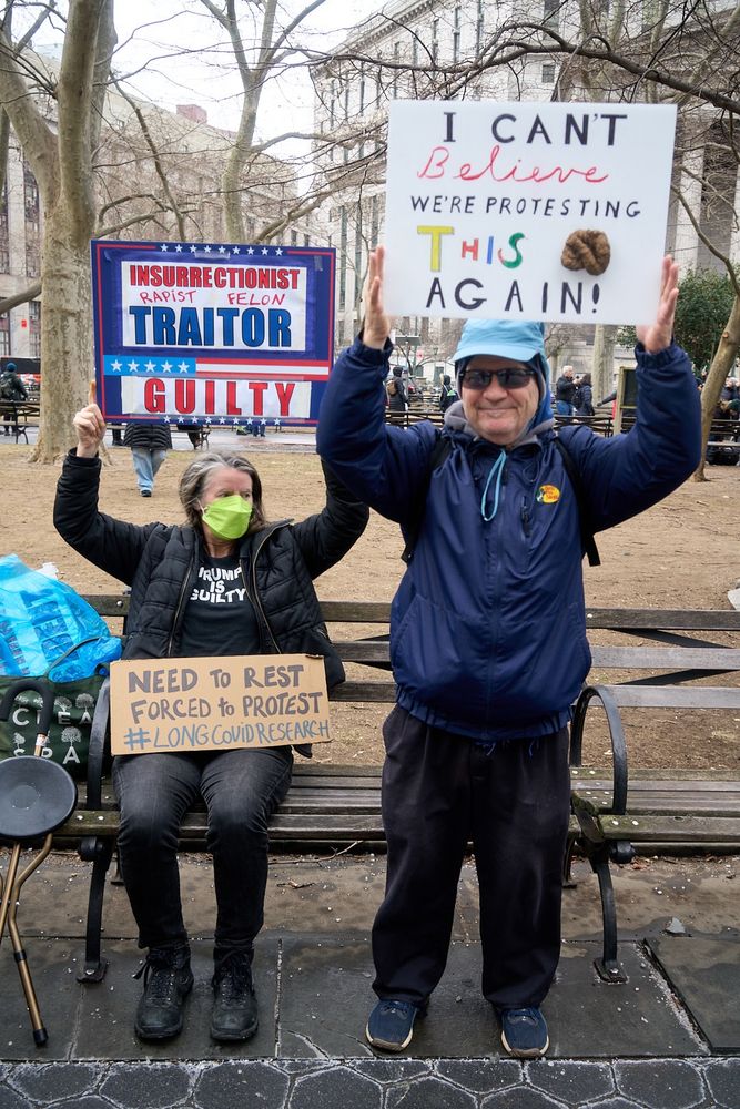 Protesters in Foley Square in NYC at the People’s March. A white lady with a lime green face mask sitting on a bench holding a sign and a white man holding a sign is standing beside her. His sign says: I CAN’T BELIEVE WE’RE PROTESTING THIS (fake turd attached to the sign) AGAIN! Her sign reads: INSURRECTIONIST RAPIST FELON TRAITOR GUILTY. Her t-shirt says TRUMP IS GUILTY. A cardboard sign sitting on her lap says NEED TO REST FORCED TO PROTEST #longcovidresearch 