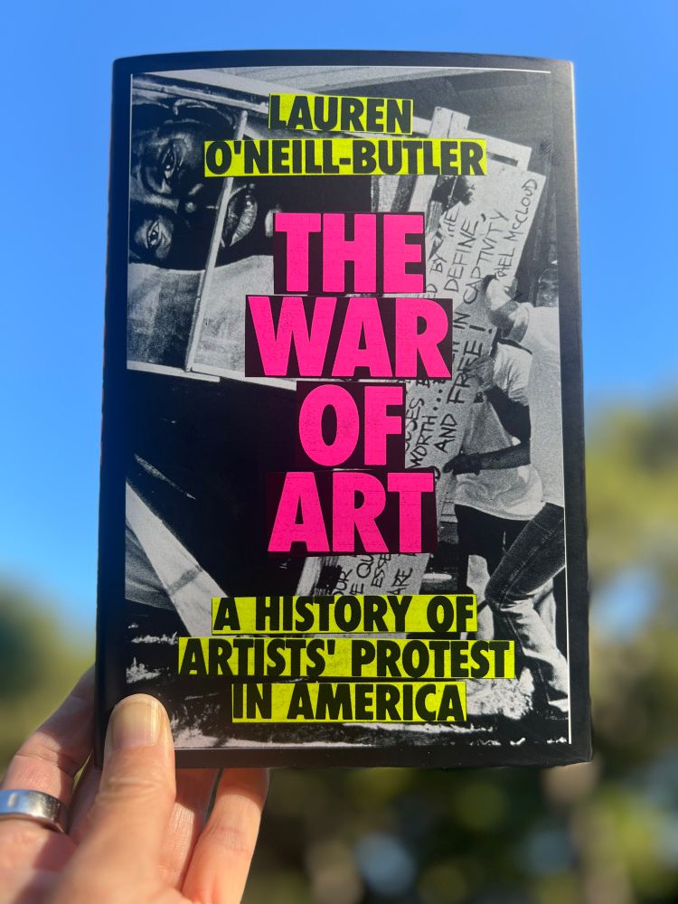 Photograph of a book taken with a phone. The book title is in large, blocky, hot-pink letters: THE WAR OF ART. The subtitle and author’s name are in black and yellow: A HISTORY OF ARTISTS’ PROTEST IN AMERICA by Lauren O’Neill Butler.

The photographer’s left hand holds up the book to catch the sun. The blue sky and trees in the background are out of focus. 

There is a silver Oura ring on the index finger to track the wearer’s heart rate & sleep data. 

The book is published by Verso. 