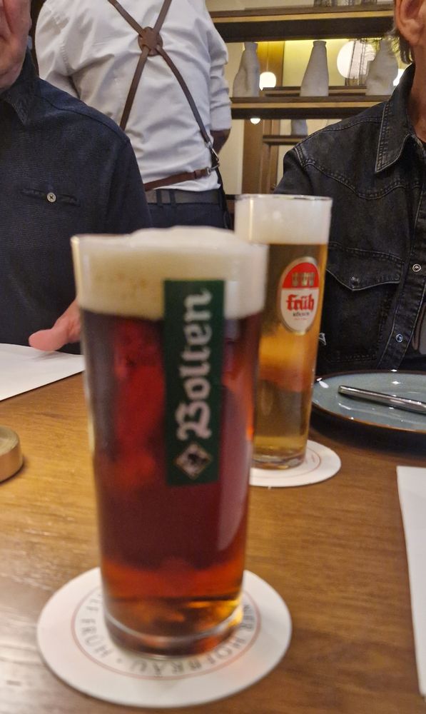 Two glasses of beer stand side by side on a table: one filled with dark Altbier in a traditional straight glass, the other with pale Kölsch in a tall, narrow glass. The scene reflects the long-standing beer rivalry between Düsseldorf and Cologne, as both styles symbolize regional identity.