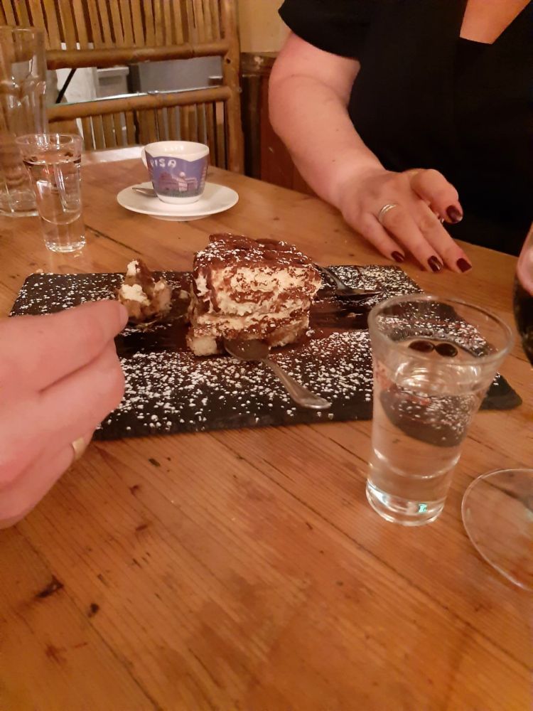 A dessert – likely tiramisu – is served on a rectangular dark plate dusted with powdered sugar. Two people are seated at a wooden table; one hand reaches in with a fork to take a bite, while the other rests casually on the table. In the foreground, there is a glass with a clear liquid, possibly grappa, and behind it, another glass and an espresso cup labeled “Pisa.” The setting appears to be a cozy, rustic restaurant.