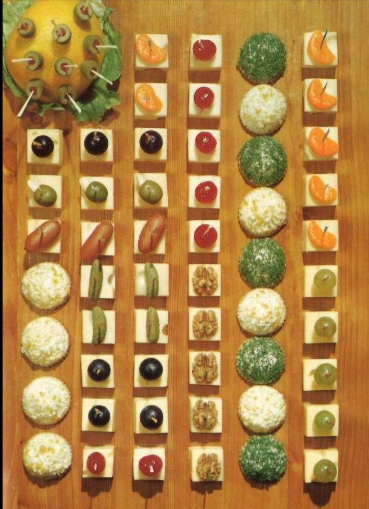 An assortment of savory and sweet canapés meticulously arranged on a wooden board. Cubes of cheese are topped with items like olives, cocktail sausages, grapes, and walnuts. On the right side are ball-shaped cheese spreads coated in herbs and coconut. In the top left, a halved grapefruit is studded with cocktail sticks.