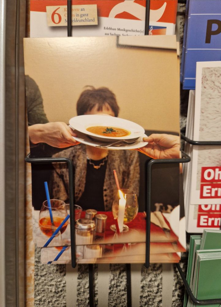 A postcard shows a dining scene: A person is being served a bowl of soup, with two hands holding the plate from opposite sides. In the background, a woman is seated at a table, but her face is mostly hidden behind the soup bowl. On the table are drinks with colorful straws, a lit candle, and condiment containers. The scene has a cozy, warm-toned atmosphere