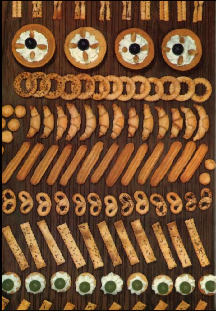 A highly symmetrical arrangement of various small baked goods on a dark wooden surface. The pastries are sorted by type into rows, including mini tarts with cream and cherries, ring-shaped cookies, crescent rolls, puff pastry sticks, pretzel-shaped cookies, and piped cream biscuits with jelly centers.