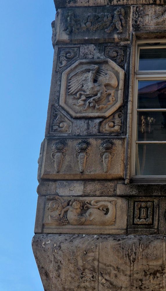 Detail of a stone relief on a building façade. It shows a large fish swallowing a reclining human figure from the lower body, with only the upper torso and head still visible. The human seems calm or resigned. Grapes and vine motifs decorate the left side. Above this relief, other sculpted elements include a stylized eagle in an octagonal frame and decorative ornaments.