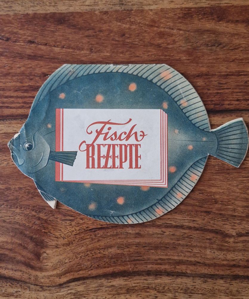 A vintage fish-shaped recipe booklet titled "Fisch Rezepte" (Fish Recipes) on a wooden surface. The booklet resembles a flatfish with a blue-green body and orange spots.