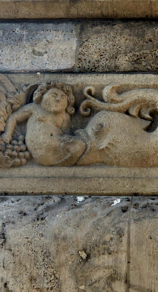 Detail of a stone relief on a building façade. It shows a large fish swallowing a reclining human figure from the lower body, with only the upper torso and head still visible. The human seems calm or resigned. Grapes and vine motifs decorate the left side. Above this relief, other sculpted elements include a stylized eagle in an octagonal frame and decorative ornaments.