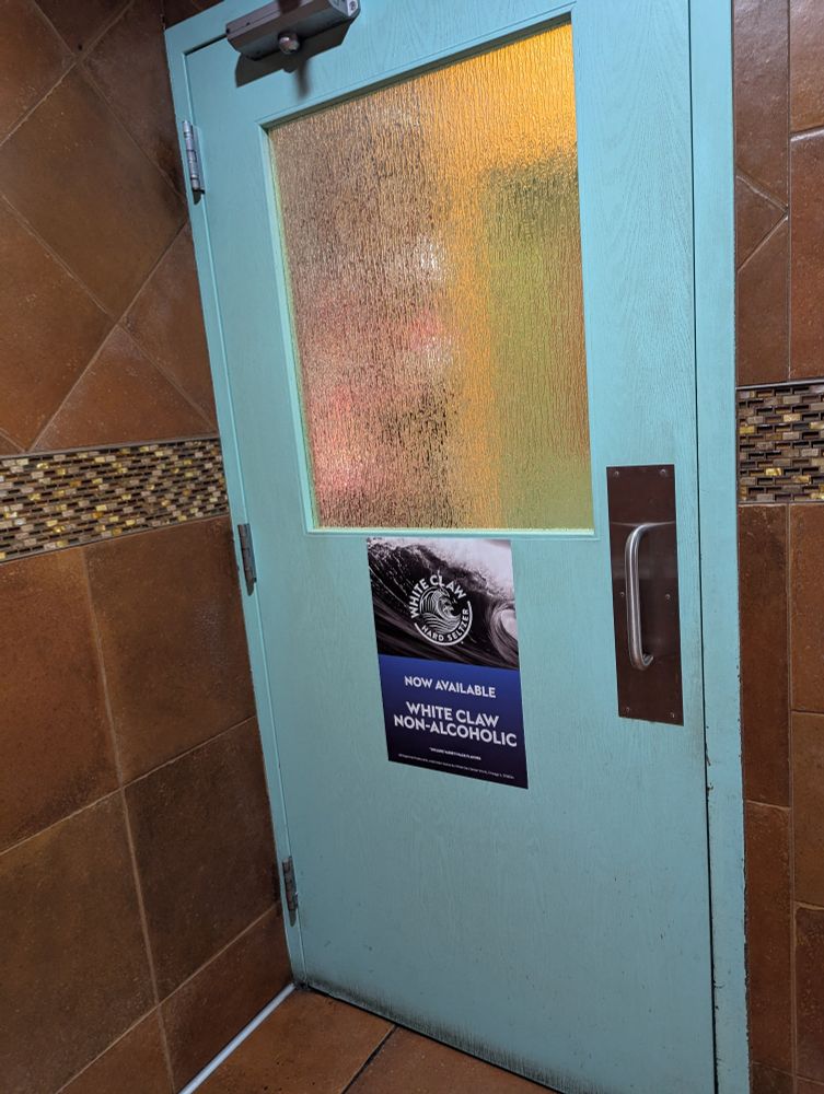 A light blue bathroom door at a restaurant with an advertisement on it for White Claw brand Non-alcoholic drink. Shit is getting seriously out of hand.