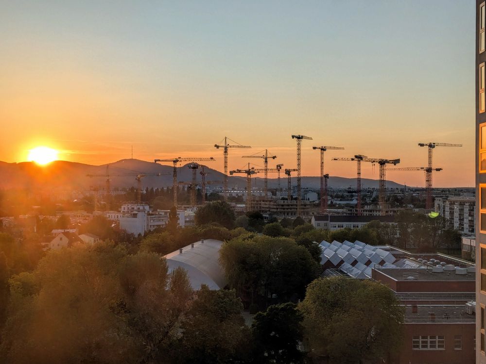 Vienna, Austria. The view is to the northwest of the sun setting behind Nussberg and Leopoldsberg (vegetated hills) with several cranes (machines) in the middle ground, and trees and buildings in the foreground.