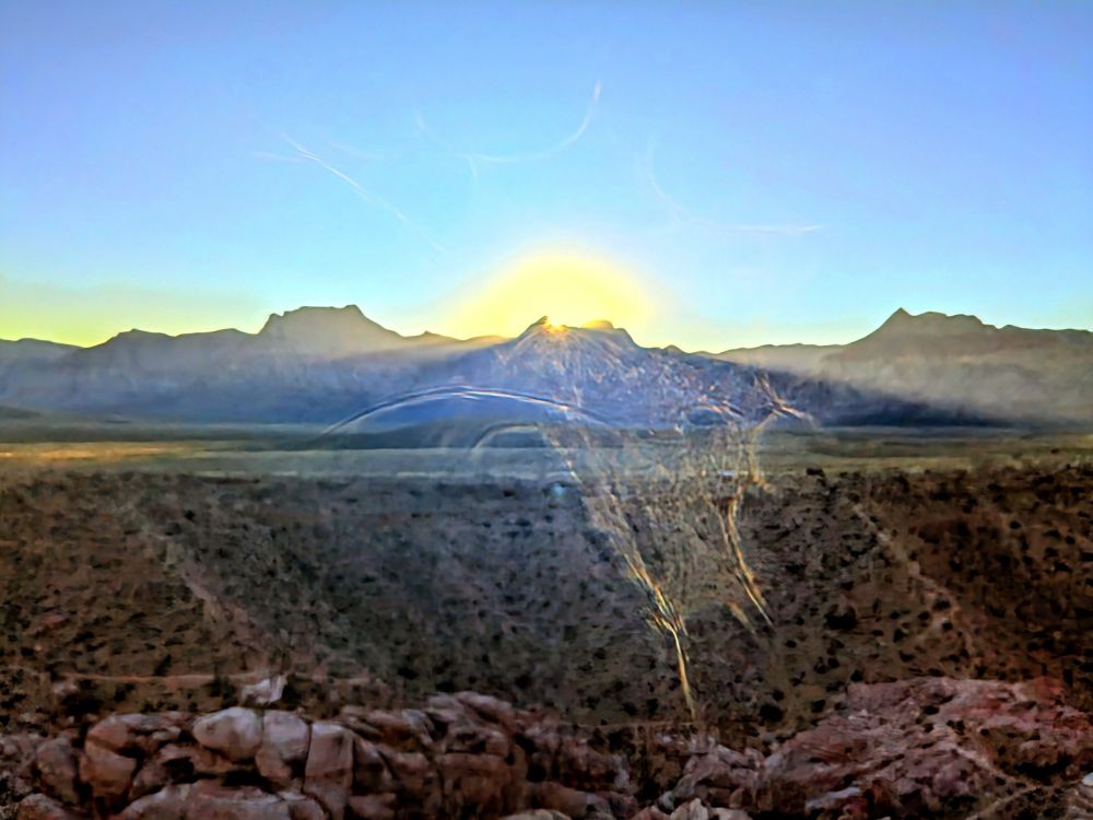 Landscape of sun going down behind craggy peaks, clear blue sky, desert vegetation, and red rocks in the foreground.