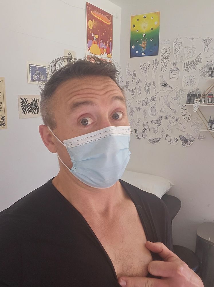 Another picture of myself. The background is the same: lots of art on the wall of various flora and fauna, as well as some more abstract pieces. You can also see some shelving still, but not as many bottles of the "Solid Ink" products.

In this picture, I'm pulling the neckline of my shirt down and to my left, to show my pectoral, which is, aside of some hair, currently bare.