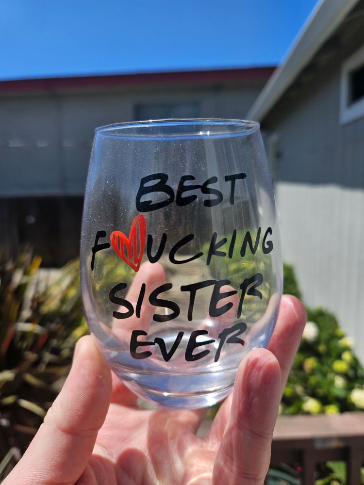 A hand holding up a stemless wine glass with "Best F❤️ucking Sister Ever" printed on it. In the blurry background is the exterior of two buildings and the greenery of what is likely a backyard or patio.