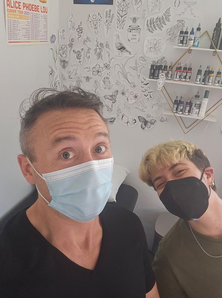 A photo of two humans wearing surgical masks. The left is me, in my standard v-neck short-sleeved shirt; eyes wide as if something is significant about the photo. 

The right human has short blonde hair and is wearing a black surgical mask, along with a thin gold chain hanging over an army green shirt. Their ears are pierced.

On the wall behind us are a number of cut out images of various flora and fauna, as well as some more abstract designs on thin sheets of paper; as well as some shelving holding small bottles of a product called "Solid Ink".