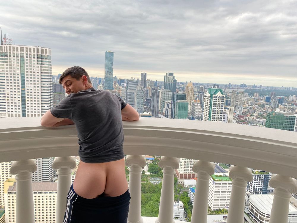 Colby Chambers leaning up against a balcony with a city skyline behind him. His shorts are pulled down to show off his ass as he invitingly looks over his shoulder 