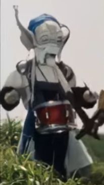 A close-up of a screencap from the TV series "Kyoryu Sentai ZyuRanger" showing Pleprechaun standing amongst the grass wearing a tin drum across his chest with drumsticks in each hand. 