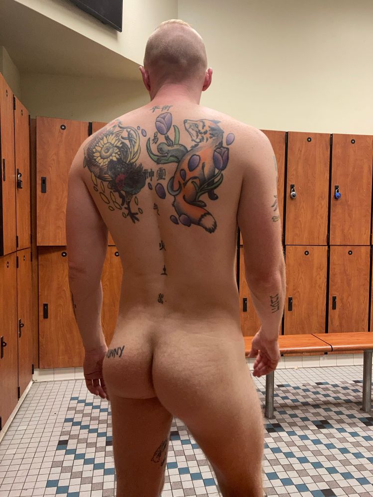 A naked man standing in a locker room revealing his butt and tattooed back.