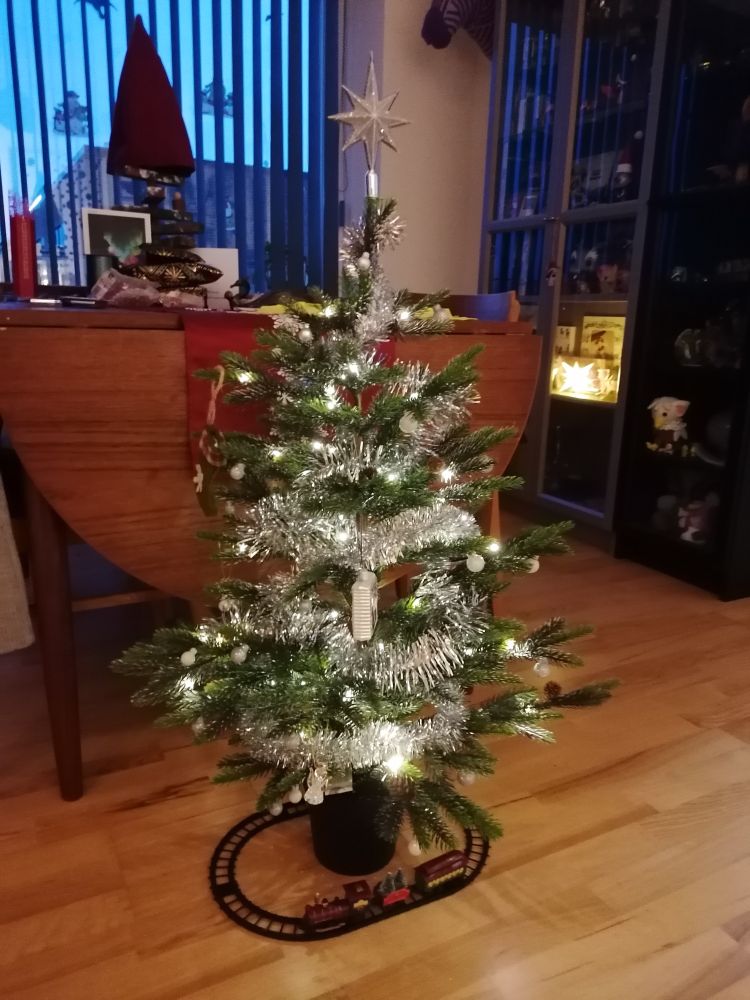 Photo of an indoor small Christmas tree, standing by a dinner table on the floor. Decorated in lights and festoon. Underneath is a small toy train on a train track going around the tree. 