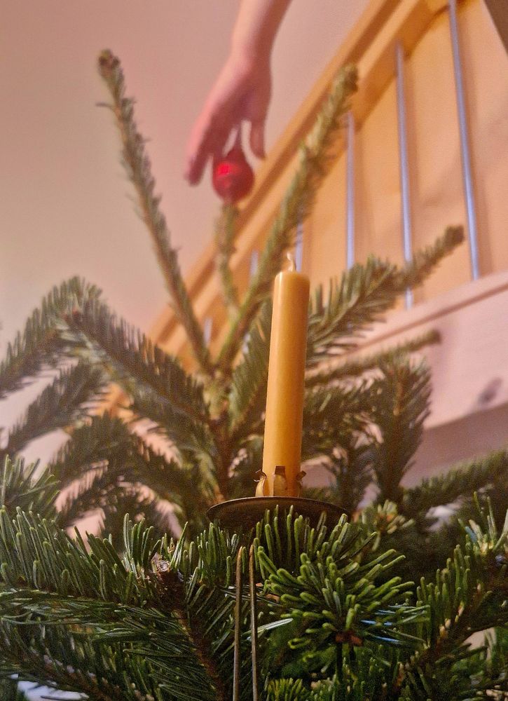Eine Kerze im Weihnachtsbaum, eine Hand setzt von oben eine rote Baumspitze auf