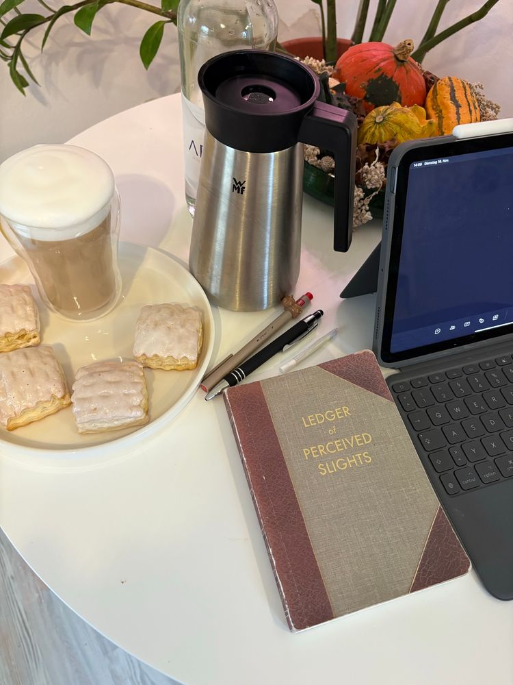 Auf einem weißen Tisch steht eine kleine Thermo Kanne unten silbern oben schwarz, ein runder weißer Teller mit vier Mini Apfelstrudel Teilchen und einem Kaffee mit Milchschaum in einem Thermo Glas, daneben liegen zwei Stifte und eine selbst gedrehte Zigarette darunter liegt ein Notizbuch mit der Aufschrift Ledger of perceived sleights am rechten Rand ist ein Teil eines Tablets mit Tastatur zusehen.