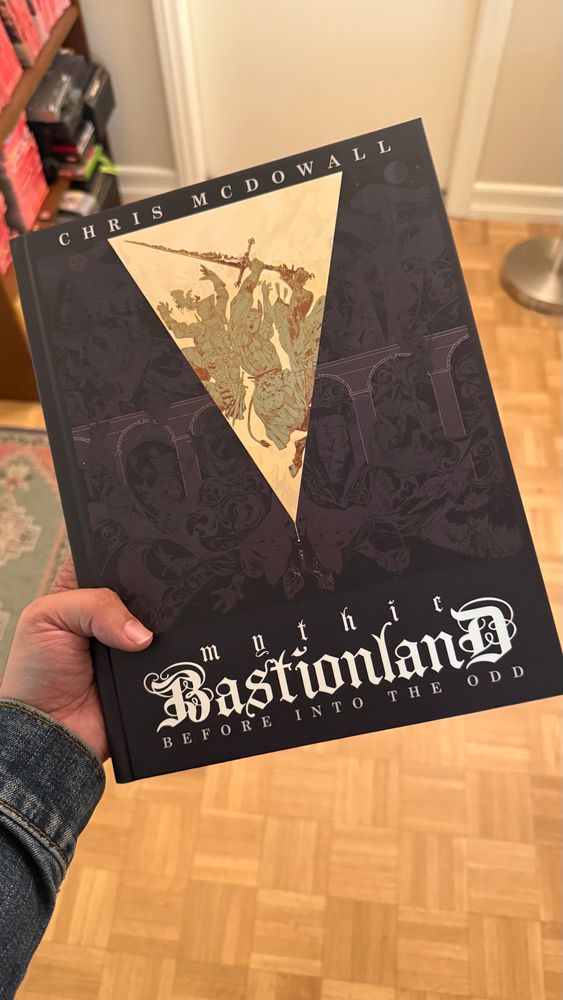 Holding up a hardbound book titled “Mythic Bastionland: Before Into the Odd” by Chris McDowell 