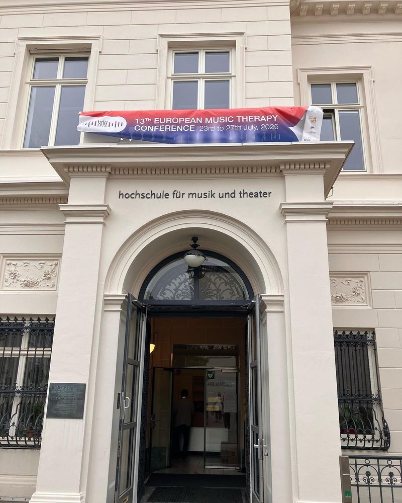 Eingangsbereich der Hochschule für Musik und Theater Hamburg mit offen stehender Glastür. Über dem Eingang hängt ein rotes Banner mit der Aufschrift „13th European Music Therapy Conference, 23rd to 27th July, 2025“.