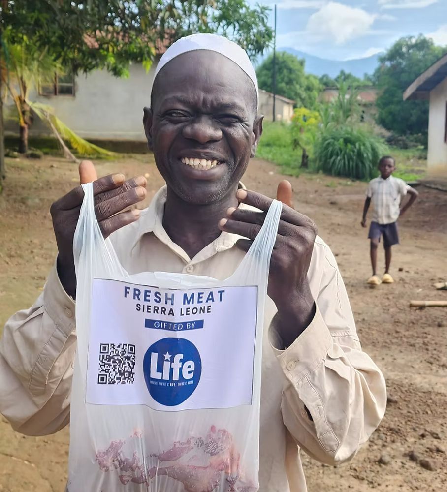 A man in Sierra Leone beams with joy while holding a plastic bag labeled “Fresh Meat – Gifted by LIFE.” The background shows a rural village setting with a child standing nearby. The image captures a moment of gratitude and dignity.