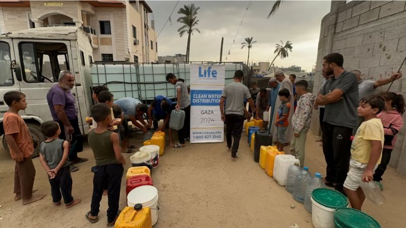 Dozens of people, including children, stand in line with yellow, white, and black water containers at a LIFE clean water distribution site in Gaza. A large water tank and banner reading "Life: Clean Water Distribution – Gaza 2024" are visible in the center. The setting is a sandy alley between buildings with palm trees in the background.