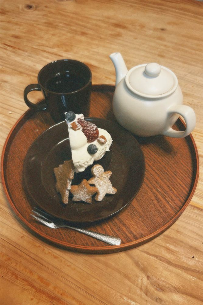 木のテーブルに置かれたケーキとお茶の写真。
丸い木のトレイの上に、茶色のマグカップと茶色の丸い皿、アイボリーのティーポット、銀のフォークが置かれている。
皿の上にはガードショコラの1ピースとツリーや星の形のクッキーが置かれている。