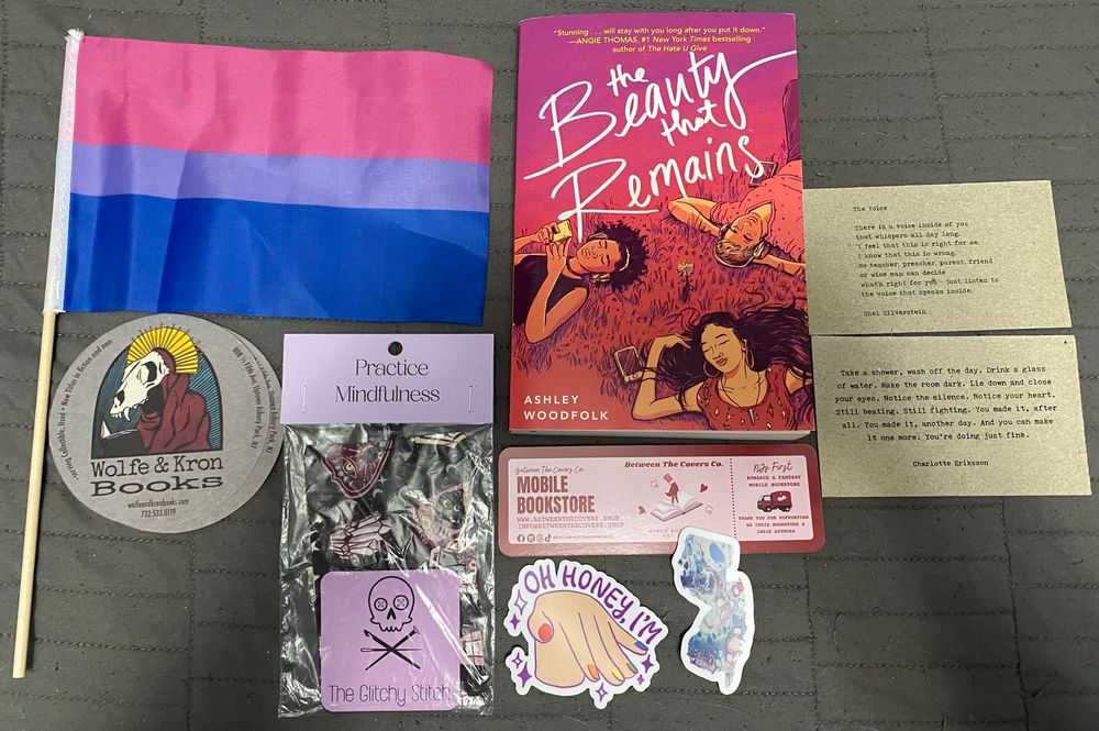 A haul from the New Jersey Bookstore Crawl. This includes a Bisexual pride flag, a Wolfe & Kron coaster, a fidget, two stickers, a bookmark, two motivational quote cards, and one book titled The Beauty That Remains by Ashley Woodfolk. 