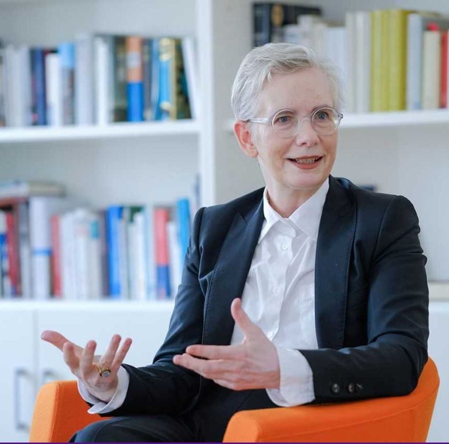 eine weiblich gelesene Person in weißer Bluse und schwarzem Jackett sitzt in einem orangefarbenen Stuhl. Darunter steht weiß auf lila: Regionalbischöfin Dr. Petra Bahr wechselt als Staatssekretärin nach Berlin
