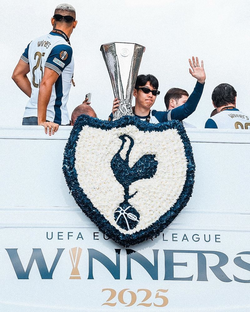 Tottenham captain Son Heung Min on top of a white, open top bus, wearing sunglasses and holding a trophy, waving to the crowd. The Spurs crest in flowers below him.