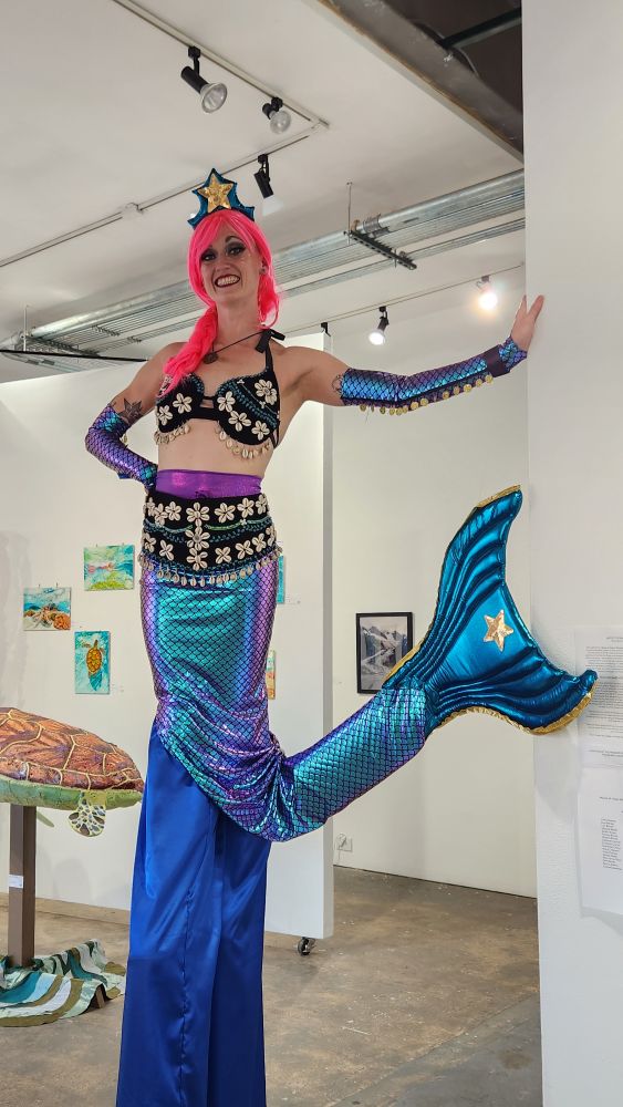 In a gallery setting, a wo.an on stilts wears a mock mermaid tail and bikini top.
