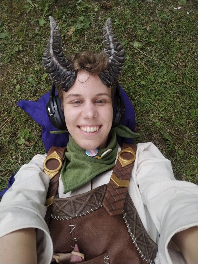 A selfie of me in Dammon (from BG3) cosplay. Laying on my back on the ground, a purple tote serving as a makeshift pillow because I'm sleepy. I'm wearing headphones instead of my pointy ears right now because I'm an autistic idiot who needs their naptime lol. The camera is raised above me and it made it seem like the viewer gets uppies from Faerûn's prettiest blacksmith.
