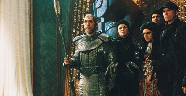 [EN]
Scene from Stargate SG-1: The Serpent’s Lair (S02E01) — Bra’tac stands at the front in silver Jaffa armor, staff weapon in hand. Behind him, from left to right, are Carter, Skaara, Teal’c, and O’Neill (with two Ls 😉), watching tensely inside a golden chamber illuminated by warm light and alien symbols.

[FR]
Scène de Stargate SG-1 : The Serpent’s Lair (S02E01) — Bra’tac se tient à l’avant, vêtu d’une armure Jaffa argentée et tenant son bâton d’arme. Derrière lui, de gauche à droite : Carter, Skaara, Teal’c et O’Neill (avec deux L 😉), observant avec tension dans une salle dorée éclairée d’une lumière chaude et de symboles extraterrestres.