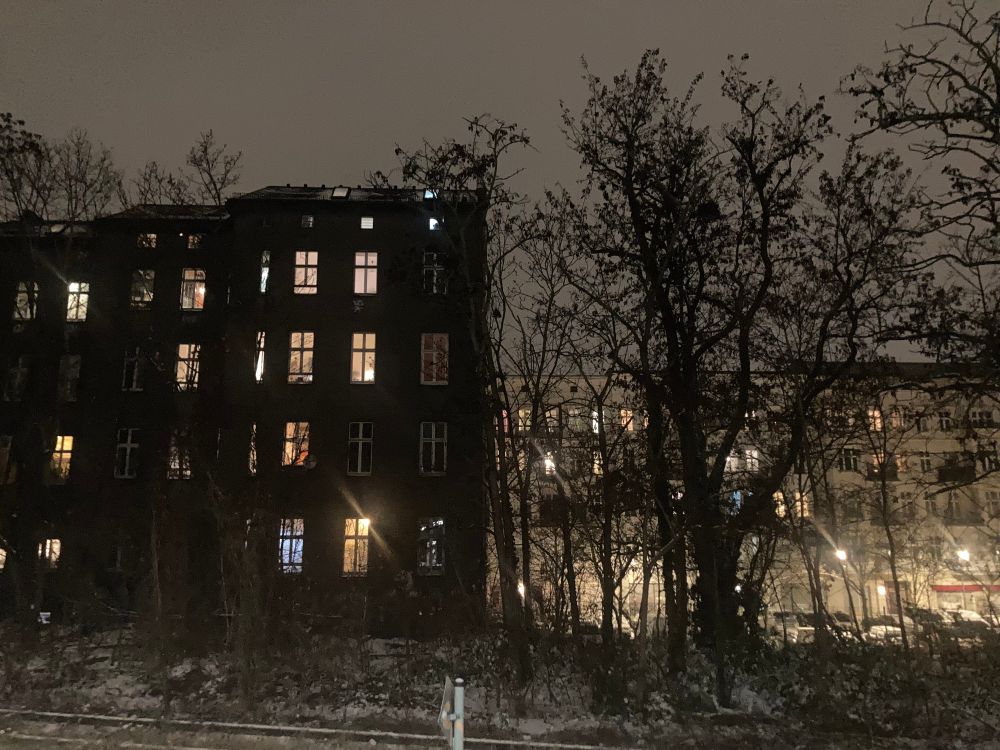 Nighttime Berlin cityscape view from Yorckstraße station.Tall dark building with warm light from multiple windows on left; tall tree silhouetted against warm streetlights on right. Sky dark grey with snow clouds