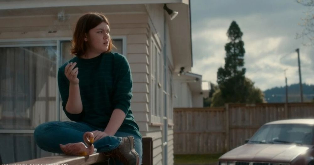 Screen capture of Shannon Berry as Dot Campbell in Amazon's The Wilds. She sits on the railing of a porch smoking a cigarette. On the railing next to her leg is a flamingo ashtray.