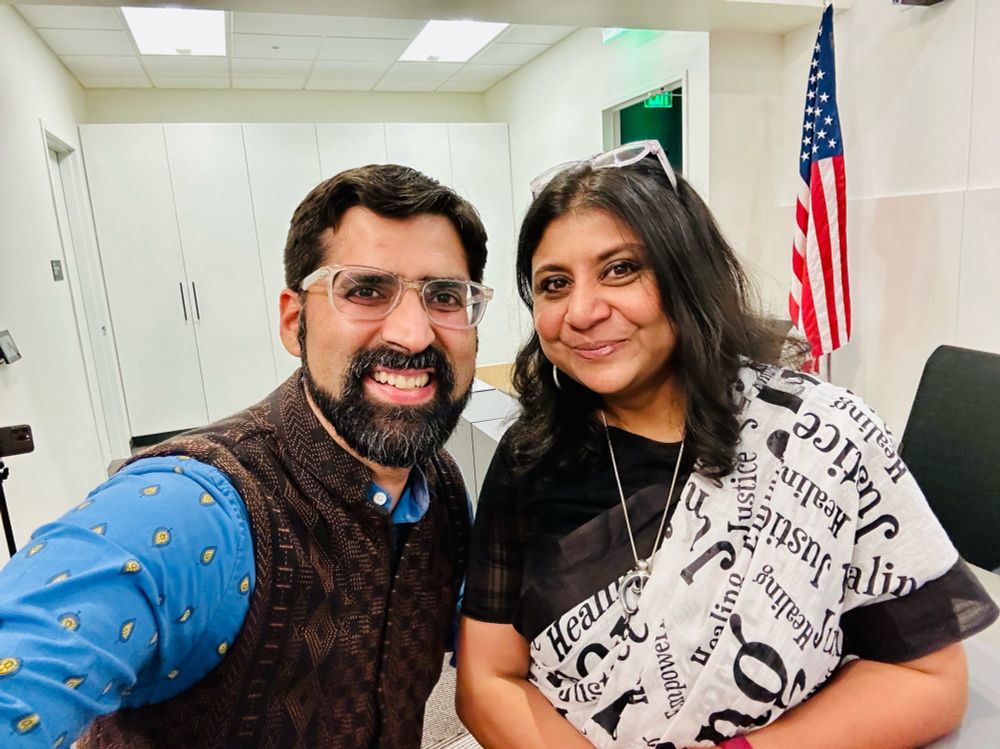 A selfie of an beaming American man in a blue shirt with a Indian American woman wearing a white sari, standing inside an office, with a US flag behind them