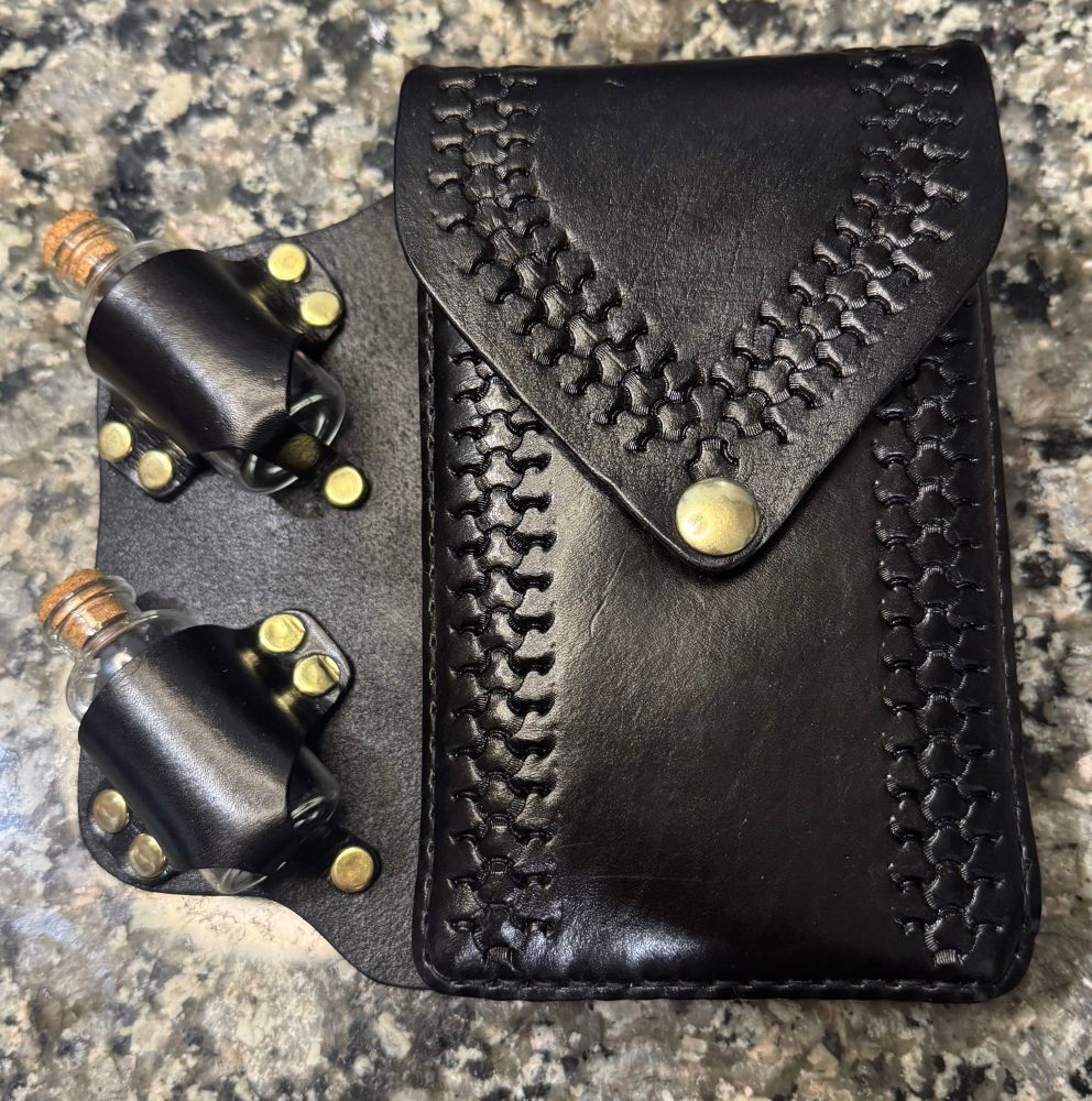 A picture of a black handmade saddlebag that holds two glass vials on the left side with a pouch on the right side that is held closed with a gold snap