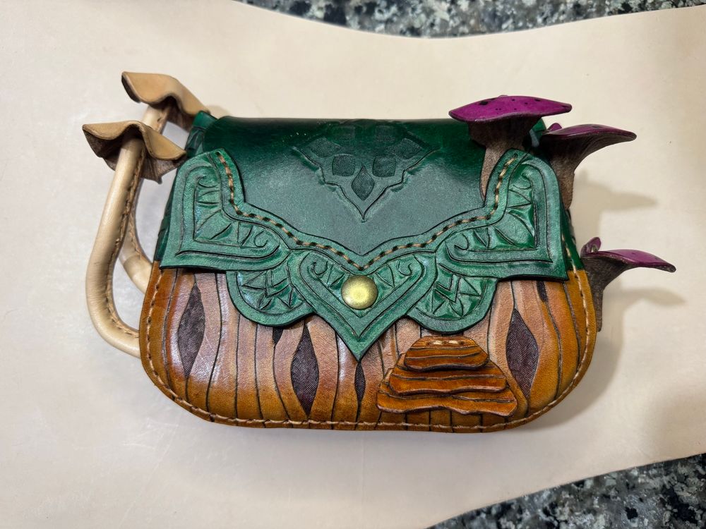 Handmade leather purse that is green on the upper half and brown on the lower half. Two tan mushrooms are sticking out of the left side and 3 purple mushrooms on the right side