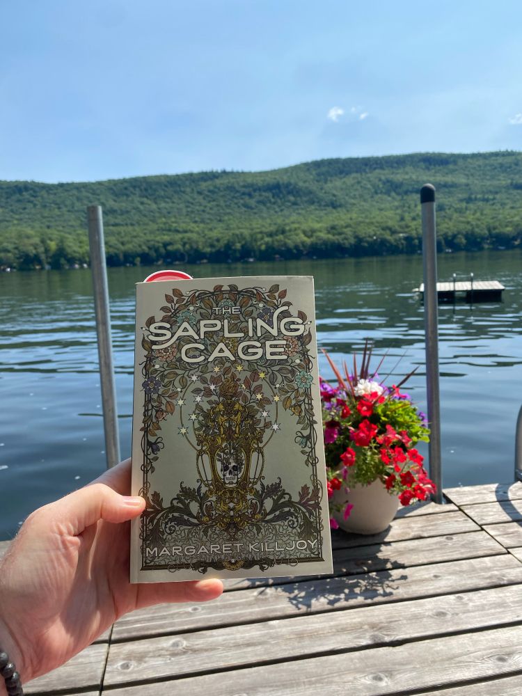 Sapling cafe by margaret killjoy with a floral design on the cover. Paperback on a dock by a mountain lake 