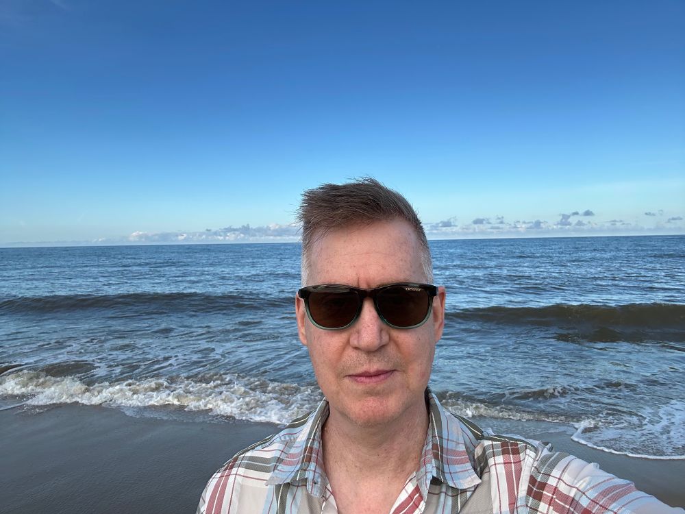 Person with short hair and sunglasses with the ocean behind them