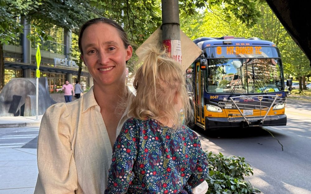 Seattle Mayoral Candidate Katie Wilson holding her daughter on a sidewalk as a King County Metro bus passes by