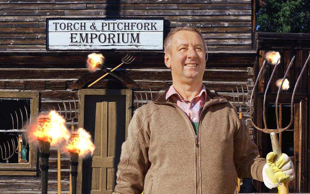 Proud business owner holding pitchfork in front of pioneer town-style building with a sign reading "Torch & Pitchfork Emporium" and assortment of pitchforks and torches outside. 