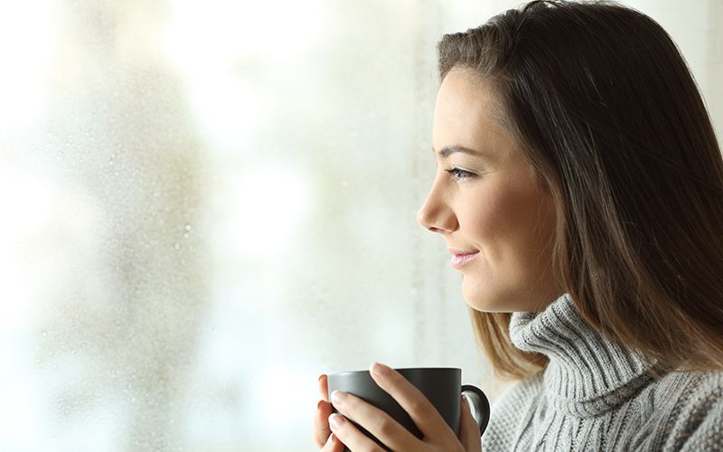 Woman in turtleneck sweater holding gray mug looking out window at gray weather with look of happy satisfaction on face