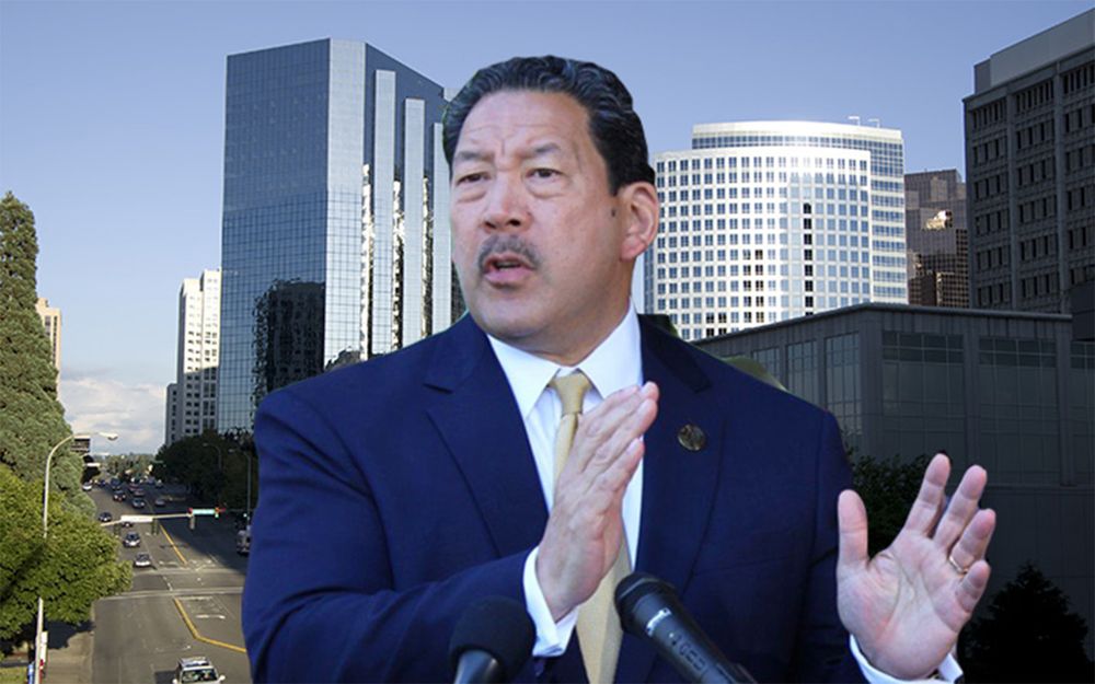 Bruce Harrell talking with stern look on his face while standing in front of bland, empty street next to bland silver office buildings in Bellevue. 