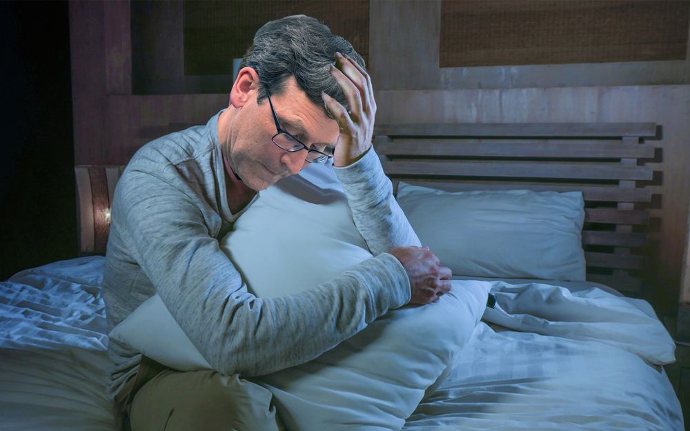 Washington State Gov. Bob Ferguson sad in dark bed hugging a pillow with his hand on his downcast face